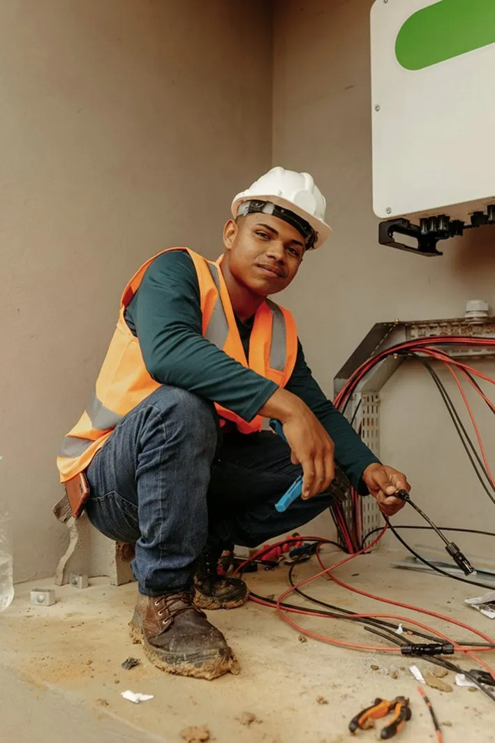 An electrician in a hard hat and hi-vis vest wiring an unfinished room
