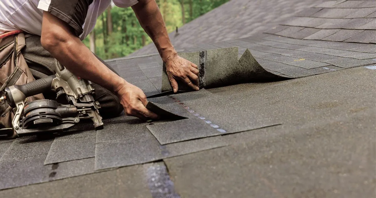 A roofer nailing down new asphalt shingles on a pitched roof