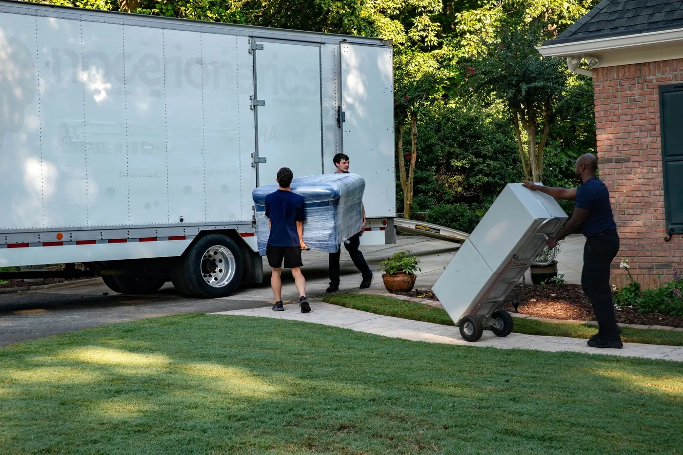 Movers loading a truck on a driveway, one wheeling an appliance up on a hand truck